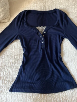 Mörkblå ribbad topp med spetsdetalj - Snygg mörkblå ribbad topp med lång ärm och knappar framtill. Toppen har en urringning med vit spetsdetalj som ger en extra touch. 