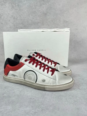 Oliver Cabell silvriga sneakers - Snygga sneakers från Oliver Cabell i silverfärgat läder med röda detaljer och röda skosnören. Skorna har svart hälparti och coola cirkeldetaljer på sidan. Klassisk låg modell med vit sula som ger en modern vibe.