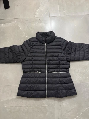 Svart pufferjacka med dragkedjor - Snygg svart pufferjacka med hög krage och raka ärmar. Jackan har två framfickor med silverfärgade dragkedjor och quiltad design. Perfekt för kalla dagar och ger en stilren look. Materialet är lätt och vadderat för extra värme.