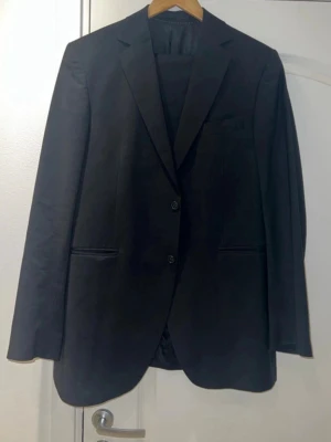 Svart klassisk kavaj - Säljer en svart kostym bestående av kavaj i storlek 52 med två knappar framtill och klassisk krage. Kavajen har raka fickor och en bröstficka, samt en stilren och tidlös design. Perfekt för dig som vill ha en enkel och snygg kavaj till olika tillfällen. Med kostymbyxor även i Tiger i storlek 50