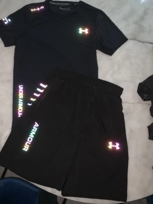 Svart träningsset från Under Armour - Snyggt svart träningsset från Under Armour med t-shirt och shorts. Båda plaggen har reflekterande loggor och text i regnbågsfärger. Materialet är lätt och andas, perfekt för träning eller gym. T-shirten har rund hals och shortsen har dragsko i midjan.
