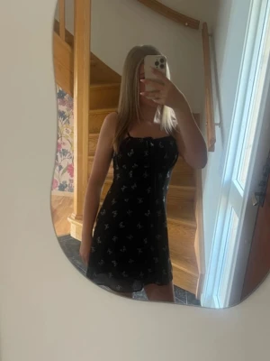 Svart kort klänning med fjärilsmönster - Söt svart kort klänning med vita fjärilar och tunna axelband. Klänningen har en lätt utsvängd passform och volangkant nedtill. Perfekt för sommarens varma dagar och enkel att styla med sneakers eller sandaler.