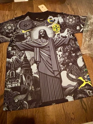 Nike Brasil grafisk t-shirt - Unik grafisk t-shirt från Nike med Brasilien-tema och stort tryck av Kristusstatyn, kyrkor och detaljerade mönster i svart, grått och gult. Rund hals och korta ärmar. Perfekt för dig som gillar streetwear och vill sticka ut.