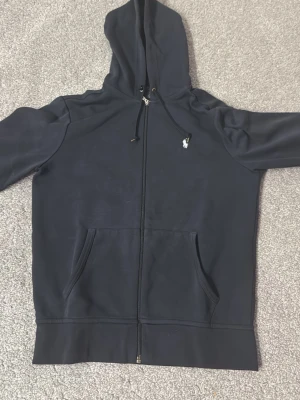 Svart hoodie från Polo Ralph Lauren - Snygg svart hoodie med dragkedja från Polo Ralph Lauren. Klassisk design med huva, dragsko och fickor framtill. Liten vit logga broderad på bröstet. Perfekt för en avslappnad och stilren look.