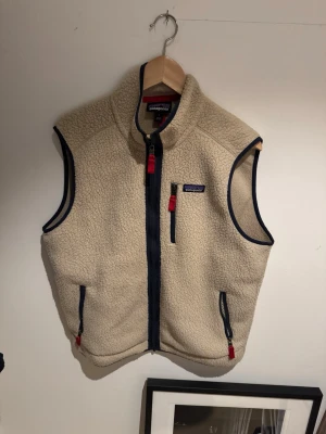 Beige fleeceväst från Patagonia - Säljer en beige fleeceväst från Patagonia med marinblå detaljer och röda dragkedjor. Västen har hög krage, tre fickor med dragkedja och är gjord i mjukt teddyfleece-material. Perfekt för lager-på-lager och riktigt skön att ha på sig.