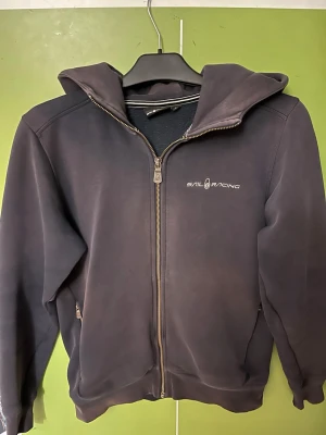 Marinblå hoodie från Sail Racing - Snygg marinblå hoodie från Sail Racing med dragkedja framtill och två fickor med dragkedja. Klassisk logga på bröstet och skön huva. Materialet känns mjukt och slitstarkt.                  Äldre modell som är använd men har skön passform.