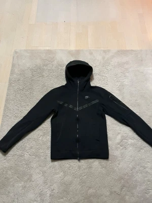 Svart Nike Tech hoodie med dragkedja XS - Snygg svart hoodie från Nike i storlek XS. Hoodien har hel dragkedja. Tillverkad i mjukt bomullsmaterial och har fickor framtill samt en extra ficka på ärmen. Skön tröja som passar alla kroppsformer.