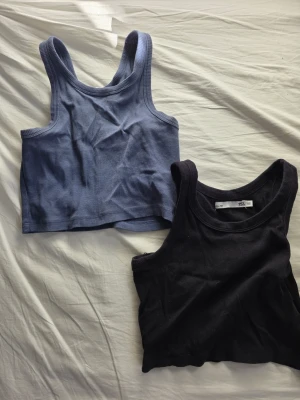 Två ribbade croppade linnetoppar - Säljer två croppade linnetoppar i ribbad bomull, en i blått och en i svart. Båda har breda axelband och rundad halsringning, perfekta för en chill och enkel stil. Passar till jeans eller shorts för en avslappnad look.