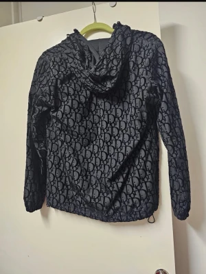 Svart vindjacka  - Snygg svart vindjacka monogrammönster över hela jackan. Jackan har huva, dragkedja framtill och två fickor med dragkedja. Tillverkad i lätt och glansigt material, perfekt för en trendig streetwear-look.