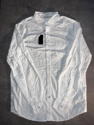 Vit skjorta med bröstficka - L - Klassisk vit skjorta från Lager157 med button-down krage och bröstficka. Skjortan har vita knappar och diskret broderad logga på fickan. Tillverkad i bomull för en skön och stilren look.