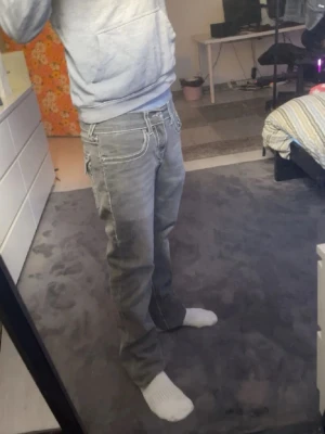 Gråa bootcut jeans med vita sömmar - Säljer ett par gråa bootcut jeans med tydliga vita kontrastsömmar och fem klassiska fickor. Jeansen har låg midja och avslappnad passform, perfekta för en casual streetstil. Materialet är denim i bomull. Snygga att matcha med hoodie eller skjorta.