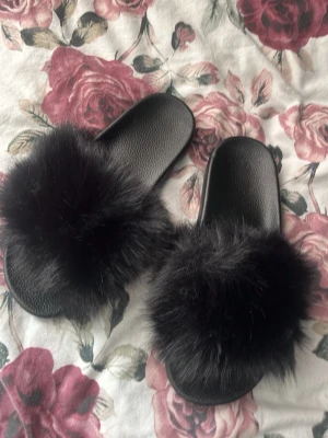 Svarta fluffiga tofflor helt nya  - Säljer ett par svarta tofflor med bred rem i mjuk fuskpäls. Sulan är i gummi och har struktur undertill för bättre grepp. Perfekta för dig som vill ha något extra mysigt och trendigt hemma. Passar till chill och avslappnade dagar.