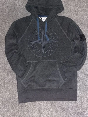 Mörkgrå hoodie från Stone Island - Snygg mörkgrå hoodie från Stone Island med broderad logga på bröstet och klassisk patch på ärmen. Hoodien har blå dragsnören och en stor magficka. Tillverkad i mjukt bomullsmaterial och har ribbade muddar vid ärmslut och nederkant.