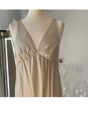 Beige kort klänning med volang - Söt beige kortklänning med v-ringad hals och breda axelband. Klänningen har en volang nedtill och är tillverkad i ett lätt, mjukt material som ger ett avslappnat intryck. Perfekt för varma dagar och enkel att styla med accessoarer.