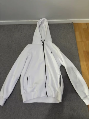 Polo Ralph lauren hoodie - Säljer en stilren vit hoodie från Polo Ralph Lauren med svart dragkedja och klassisk huva med dragsko. Hoodien har två fickor framtill och den ikoniska lilla Polo-loggan på bröstet. Perfekt för en clean och avslappnad look.