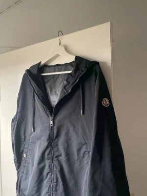Mörkblå windbreaker från Moncler - Snygg mörkblå vindjacka från Moncler med dragkedja framtill. Jackan har Monclers klassiska logotyp på ärmen och är tillverkad i ett lätt, vindtätt material som passar perfekt för blåsiga dagar. Pris kan diskuteras vid snabb affär!
