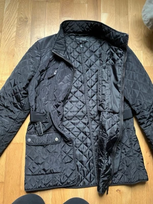 Svart quiltad jacka  - Snygg svart quiltad jacka  med bälte i midjan och flera fickor framtill. Jackan har dragkedja och knappar, lång ärm och en klassisk krage. Perfekt för kyligare dagar och ger en stilren look.