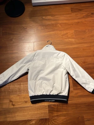 Vit vindjacka  - Vit vindjacka med svart dragkedja och svarta muddar. Broderad text och detaljer på bröstet. Jackan har ståkrage, två fickor fram och elastiska muddar vid ärmslut och midja. Perfekt för blåsiga dagar och snygg till jeans.