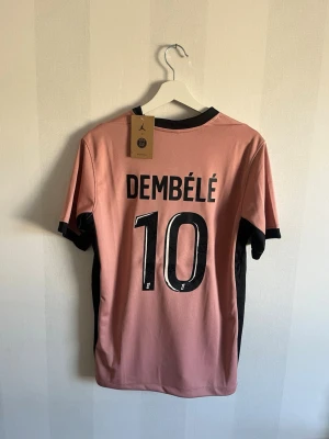 PSG rosa matchtröja Dembélé #10 - Snygg rosa PSG matchtröja med svarta detaljer, Dembélé och nummer 10 på ryggen. Framsidan har Qatar Airways-logga, PSG-märke och Jordan-logga. Korta ärmar och sportig passform, perfekt för fotboll eller streetwear.
