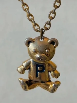 Guldfärgat halsband med nalleberlock - Äkta 90tal vintage. Unikt halsband med en guldfärgad kedja och en söt nalleberlock. Nallen har en tröja med ett blått 'P' på bröstet och detaljerade armar och ben. Perfekt accessoar för dig som gillar personliga smycken och lekfull design.