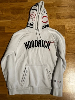 Vit hoodie från Hoodrich med tryck - Vit hoodie från Hoodrich med svart och rött tryck på bröstet och textdetaljer på huvan och ärmen. Stor känguruficka framtill och tryckt text på ryggen. Oversized passform och mjukt material, perfekt för en streetwear-look.