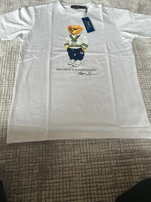 Vit Polo Bear t-shirt från Ralph Lauren - Vit t-shirt från Ralph Lauren med ikoniska Polo Bear-trycket framtill. Björnen har på sig en vit tröja med RLTC-tryck och blå byxor. T-shirten har rund hals och korta ärmar. Tillverkad i mjuk bomull, perfekt för en avslappnad och stilren look.