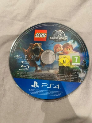 LEGO Jurassic World PS4 spel - Säljer spelet LEGO Jurassic World till PlayStation 4. Skivan är rund och har färgglad etikett med dinosaurier och LEGO-karaktärer. Märkt med PS4-logga och PEGI 7. Perfekt för dig som gillar äventyr och LEGO-spel.