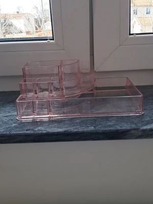 Rosa sminkorganiserare i plast - Transparent rosa sminkorganiserare med flera fack i olika storlekar för förvaring av sminkborstar, pennor och andra skönhetsverktyg. Tillverkad i tålig plast och har en modern, stilren design som passar perfekt på sminkbordet. Har 2 sprickor. 