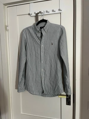 Randig skjorta från Polo Ralph Lauren - Klassisk vit och ljusgrön randig skjorta från Polo Ralph Lauren med button-down krage och broderad logga på bröstet. Skjortan har långa ärmar och knäppning framtill. Perfekt för en clean och stilren look.