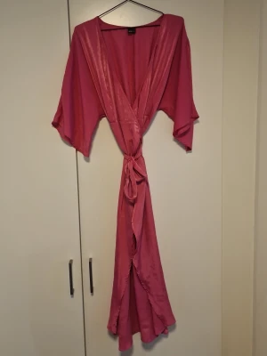 Mörkrosa omlottklänning  - Snygg mörkrosa omlottklänning med vid ärmar och knytskärp i midjan. Tillverkad i glansigt satinliknande material som känns mjukt mot huden. Perfekt för att chilla hemma i och ger en lyxig känsla.