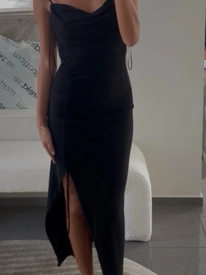 Svart långklänning med slits - Snygg svart långklänning med tunna axelband och draperad urringning. Klänningen har en hög slits framtill och figurnära passform som framhäver siluetten. Perfekt för dig som vill ha en elegant och stilren look.