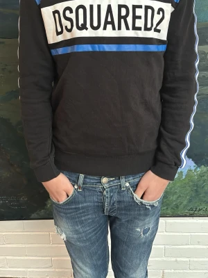 Dsquared2 - Svart långärmad t-shirt från Dsquared2 med vit och blå detalj på bröstet och blå/vita ränder längs ärmarna. Stor logga framtill. Skön passform och ribbade muddar. Perfekt för dig som gillar streetwear och vill sticka ut.