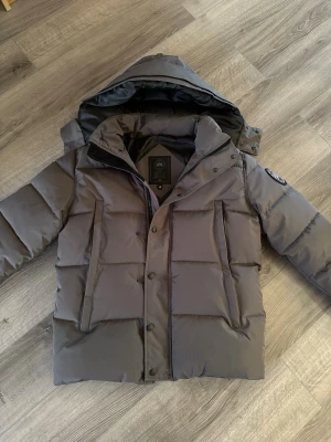 Grå pufferjacka med huva Canada Goose - Säljer en grå pufferjacka från Canada Goose med stor huva och diskret logotyp på ärmen. Jackan har dragkedja och knappar framtill, två stora vertikala fickor och quiltad design. Perfekt för kalla vinterdagar och riktigt snygg streetstil.