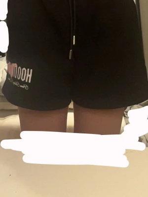 Svarta shorts från Hoodrich - Svarta shorts från Hoodrich med tryckt logga i vitt och rosa på vänster ben. Modellen har elastisk midja med snörning och är tillverkad i mjuk bomull, perfekta för chill eller träning. Klassisk loose fit och enkel design.