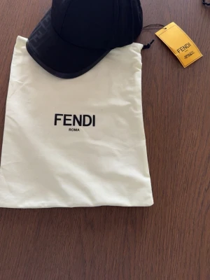 Svart keps från Fendi - Snygg svart keps från Fendi med diskret logomönster på skärmen. Klassisk böjd skärm och justerbar passform. Kepsen kommer med original dustbag och tagg. Perfekt accessoar för en trendig och exklusiv look. Den är helt ny aldrig använt