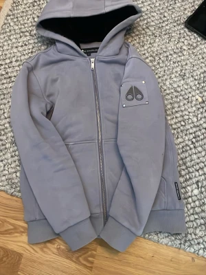 Ljuslila hoodie från Moose Knuckles - Snygg ljuslila hoodie från Moose Knuckles med dragkedja framtill och huva. Har ett metallmärke med logga på ena ärmen och ribbade muddar vid ärmslut och nederkant. Perfekt för en chill och stilren look. Och om ni har några funderingar eller något så är det Bara att höra av sig 