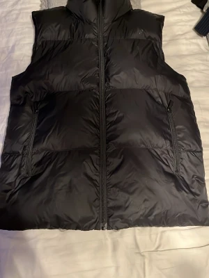 Svart dunväst från Dsquared2 - Snygg svart dunväst från Dsquared2 med hög krage och dragkedja framtill. Västen har två fickor med dragkedja och logga tryckt i vitt på kragen baktill. Perfekt för lager på lager och streetwear-stil. Den är 14 men den passar s