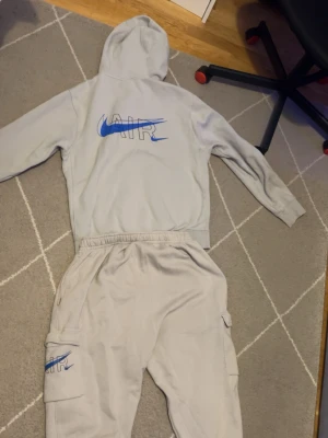 Nike Air grått mjukisset - Säljer ett grått mjukisset från Nike Air med blå logga och text på både hoodie och byxa. Hoodien har huva med dragsko och byxorna har resår i midjan samt fickor på sidorna. Perfekt för en avslappnad och sportig stil.