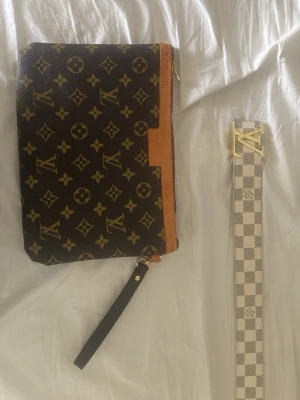 Monogram Clutch - Snygg clutch från i klassiskt brunt monogrammönster med beige och guldiga detaljer. Väskan har en avtagbar handledsrem i svart läder och dragkedja upptill. Perfekt accessoar för dig som gillar lyxig och tidlös design.