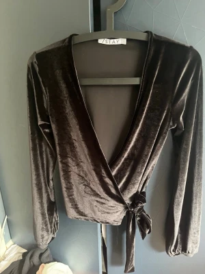 Svart omlottopp i sammet från Stay - Säljer en svart omlottopp från Stay i mjuk sammet. Toppen har långa ärmar, djup v-ringning och knyt i sidan som ger en snygg siluett. Perfekt för dig som gillar trendiga och stilrena plagg med lite extra lyxig känsla.