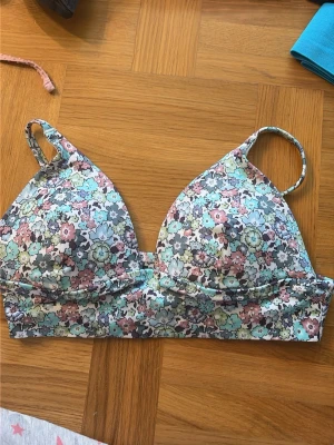 Blommig bikiniöverdel i pastellfärger - Söt bikiniöverdel med blommigt mönster i pastelliga toner av blått, rosa, grönt och vitt. Triangelformade kupor med justerbara axelband. Perfekt för stranddagar och sommarhäng. Säljer då den är för liten. Aldrig använd. 