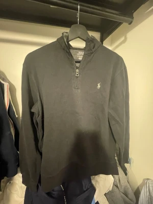 Mörkblå quarter zip från Polo Ralph Lauren - Svart långärmad tröja från Polo Ralph Lauren med klassisk quarter zip och broderad logga på bröstet. Tröjan har hög krage och ribbade muddar vid ärmslut och nederkant. Perfekt för en avslappnad men ändå stilren look.