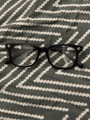 Svarta solglasögon från Chrome Hearts - Snygga svarta solglasögon från Chrome Hearts med klassisk rak form och tydliga loggor på skalmarna. Glasögonen har en stilren och modern look som passar perfekt till streetwear och trendiga outfits.