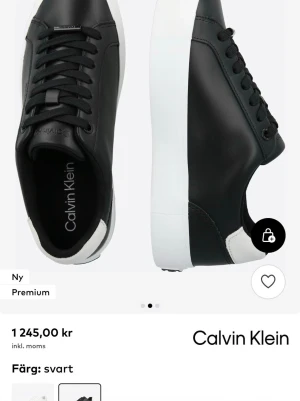 Svarta sneakers från Calvin Klein - Stilrena svarta sneakers från Calvin Klein med vit sula och diskret logga på sidan. Skorna har lågt skaft, svarta skosnören och en kontrasterande vit detalj vid hälen. Tillverkade i slätt syntetmaterial för en clean look.