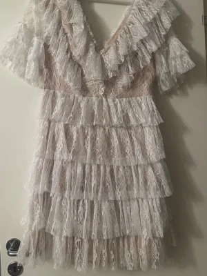 Vit spetsklänning med volanger - Supersöt kort vit klänning i spets med flera lager volanger och v-ringning både fram och bak. Klänningen har korta ärmar och ett romantiskt uttryck, perfekt för dig som vill sticka ut. Materialet är lätt och luftigt, vilket gör den skön att bära.