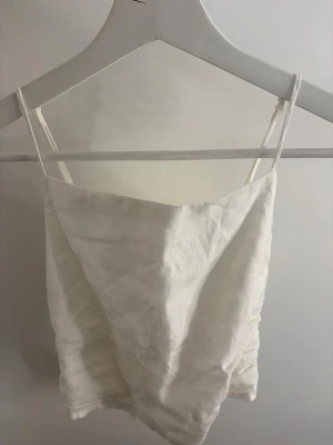 Vit linnetopp med knyt i ryggen - Säljer en vit linnetopp med tunna justerbara axelband. Den har en snygg öppen rygg med knytning som går att justera själv. Tröjan är använd endast 2 gånger. 