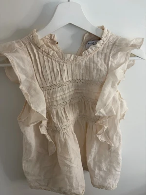 Krämvit blus med volanger Holly & Whyte - Söt krämvit blus från Holly & Whyte by Lindex med volangdetaljer längs ärmhålen o nedåt, och fina spetsinslag.