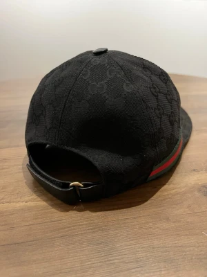 Äkta Gucci med GG-mönster - Väldigt lite använd. Snygg svart keps från Gucci med klassiskt GG-mönster i tyget. Kepsen har en böjd skärm och justerbart spänne bak i läder. På sidan finns den ikoniska gröna och röda randen. Perfekt accessoar för dig som gillar exklusiv streetwear.