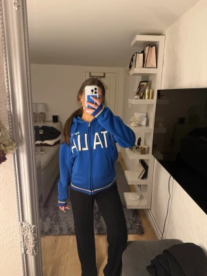 Blå Italia hoodie med dragkedja - Snygg blå hoodie med dragkedja och huva från Italia. Stora vita bokstäver framtill, ribbade muddar och fickor på sidorna. Perfekt för dig som gillar sportig och avslappnad stil. Tröjan har även Italiens flagga på bröstet och vita detaljer vid ärmslut och midja. Köpt i Rom