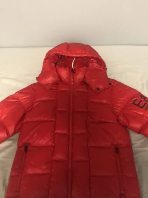 Röd dunjacka EA7 Emporio Armani - Snygg röd dunjacka från EA7 Emporio Armani med stor logga på ärmen. Jackan har huva, dragkedja framtill och två sidofickor med dragkedja. Puffig modell som håller dig varm under kalla dagar. Perfekt för dig som vill sticka ut i vinter.